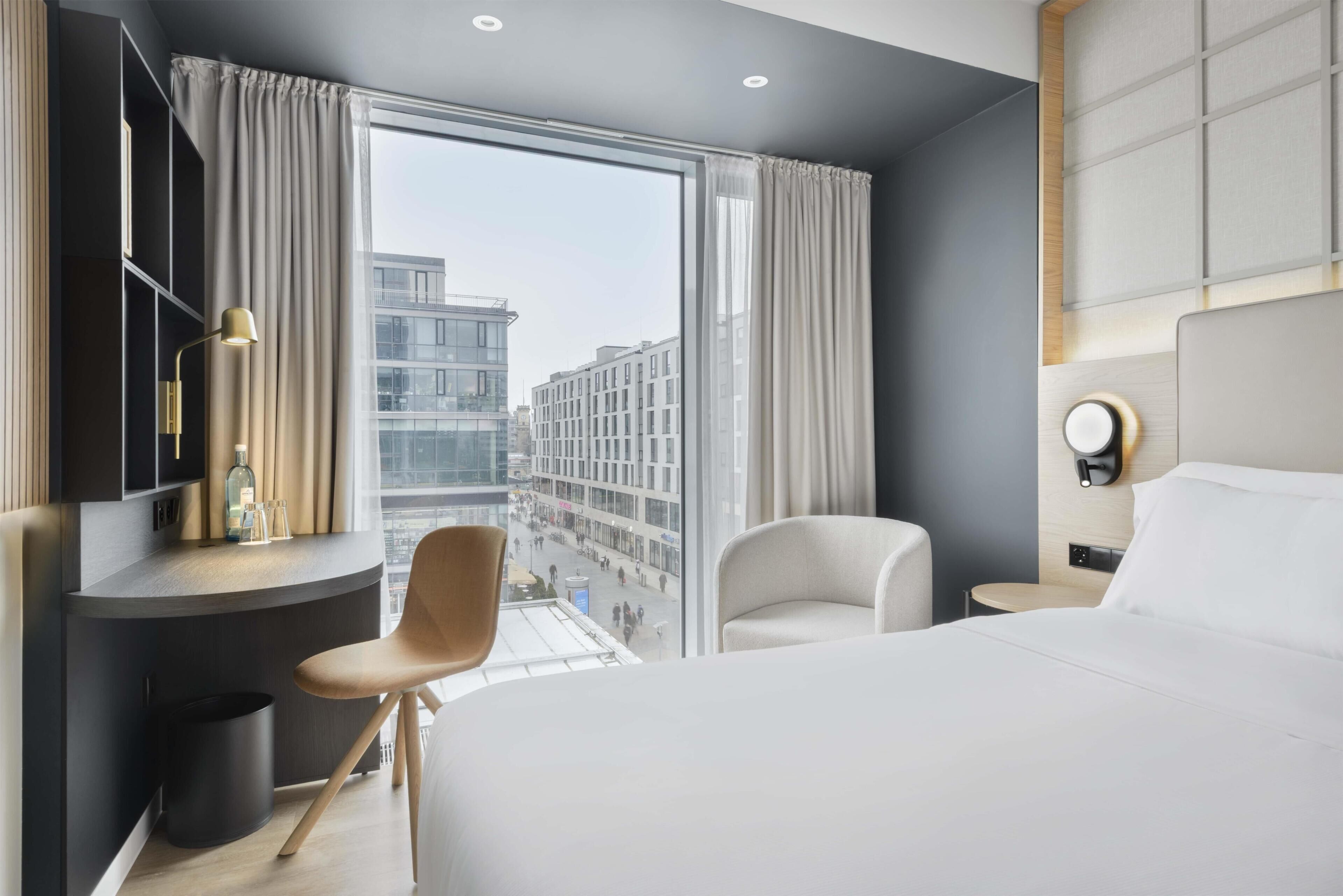 A modern hotel room with a large bed, a desk with a chair, a bedside table, a lamp, and a view of the cityscape.