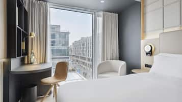 A modern hotel room with a large bed, a desk with a chair, a bedside table, a lamp, and a view of the cityscape.