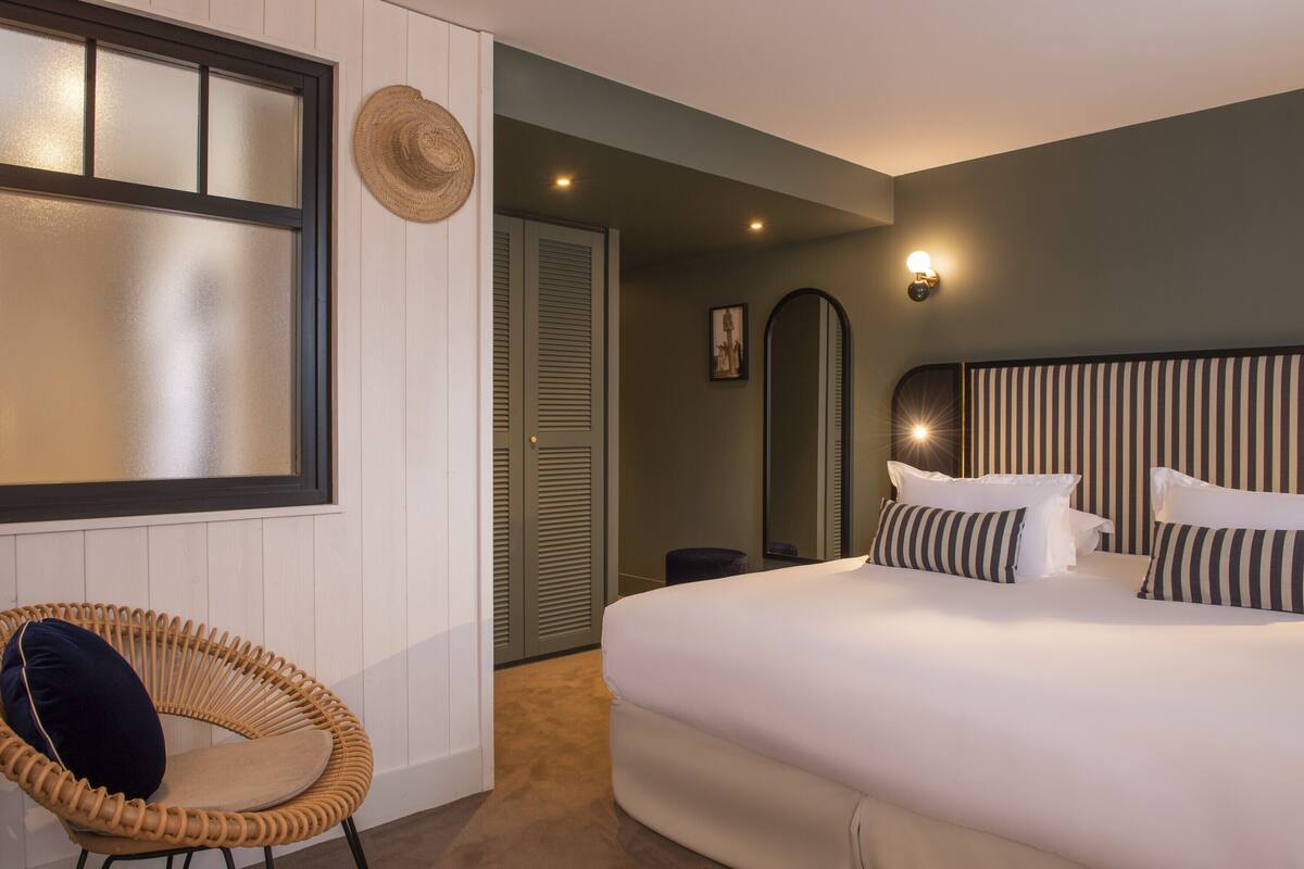 A hotel room with a large bed, a wicker chair, a mirror, and a wall-mounted light fixture.