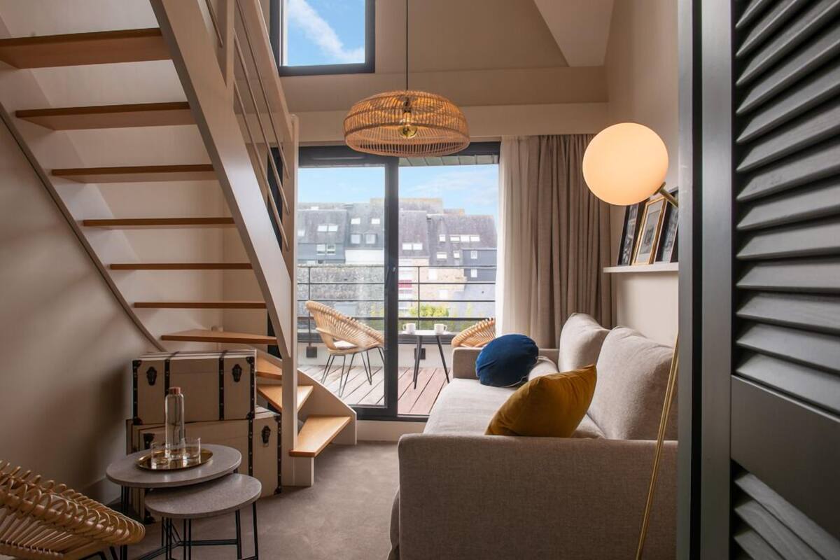 A modern living room with a staircase, a sofa with cushions, a round table, wicker chairs, and a view through a large window.