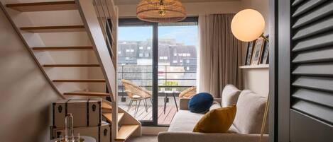 Una sala moderna con una escalera, un sofá con cojines, una mesa redonda, sillas de mimbre y vistas a través de un ventanal.