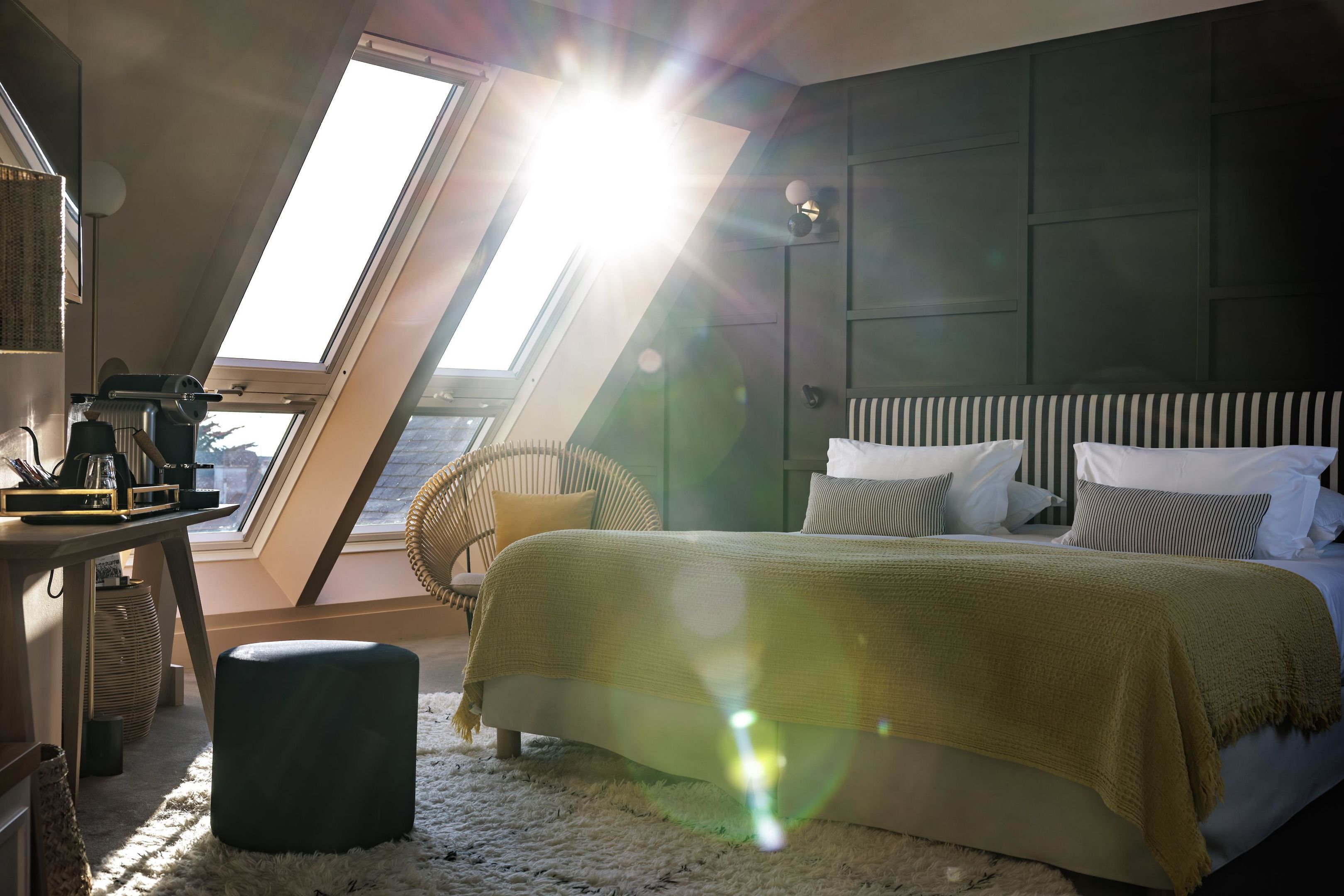Un dormitorio con tragaluces, una cama con ropa de cama a rayas, una silla y una mesita de noche con lámpara.
