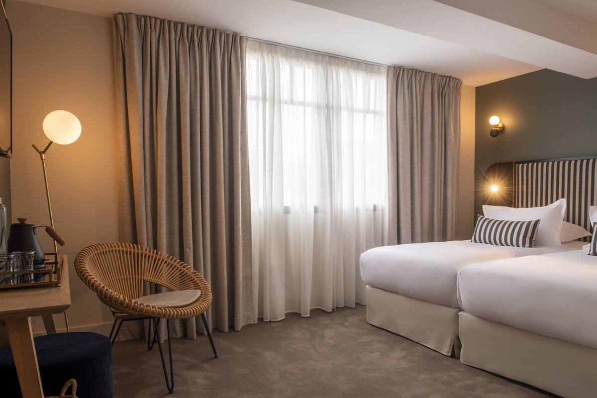 A hotel room with a large bed, a chair, a desk with a lamp, and curtains.