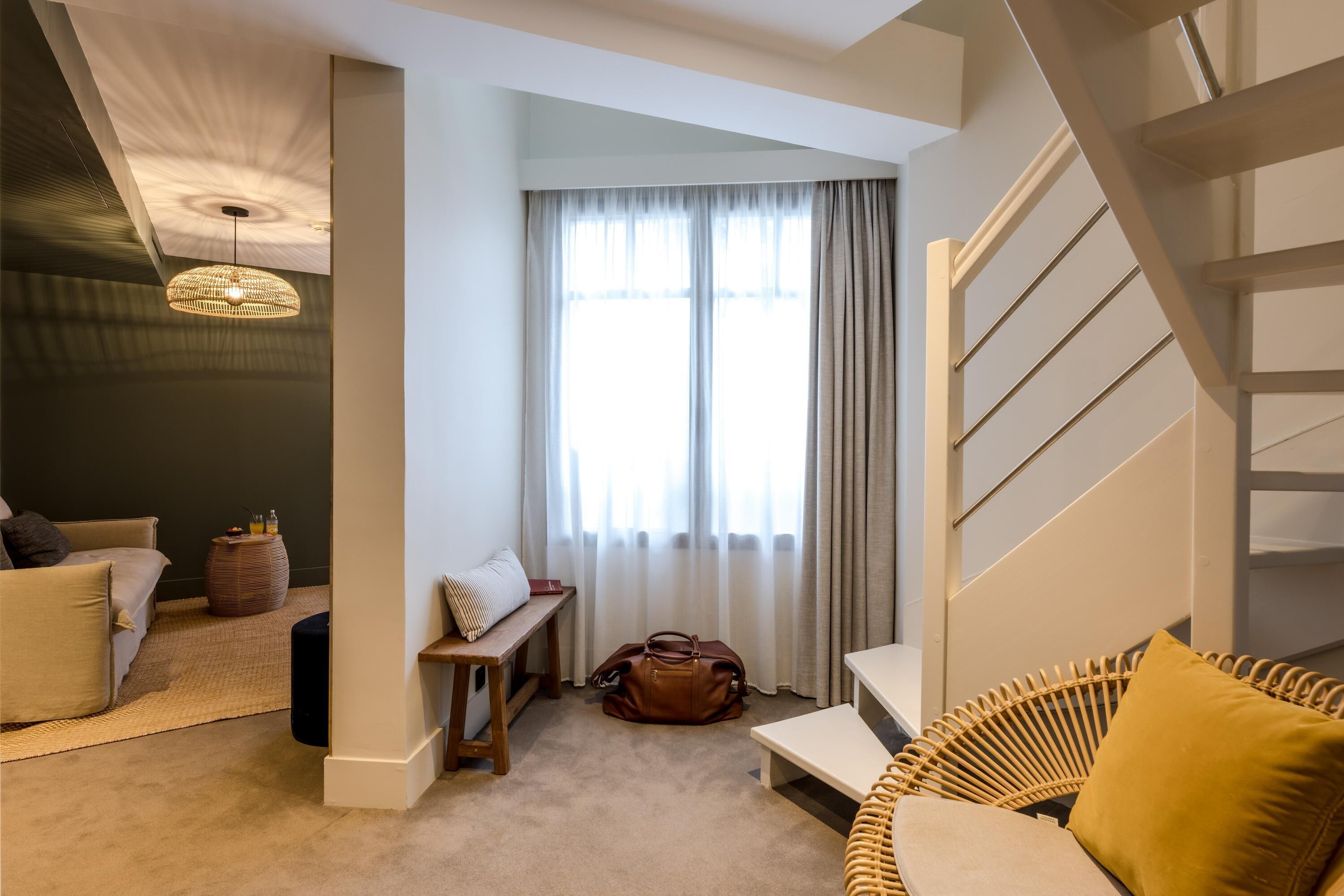 En moderne stue med sofa, bænk, fletstol, trappe og et stort vindue med gardiner.
