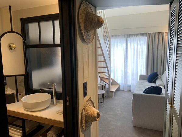A modern hotel room with a large mirror, a white sink, a round bowl, a wooden door, a staircase, a sofa with blue pillows, and a window with curtains.