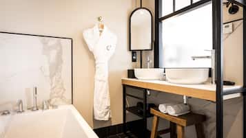 A modern bathroom with a marble wall, a white bathtub, a wooden vanity with a sink, a mirror, and a robe hanging on the wall.