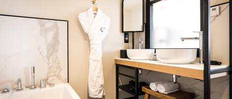A modern bathroom with a marble wall, a white bathtub, a wooden vanity with a sink, a mirror, and a robe hanging on the wall.