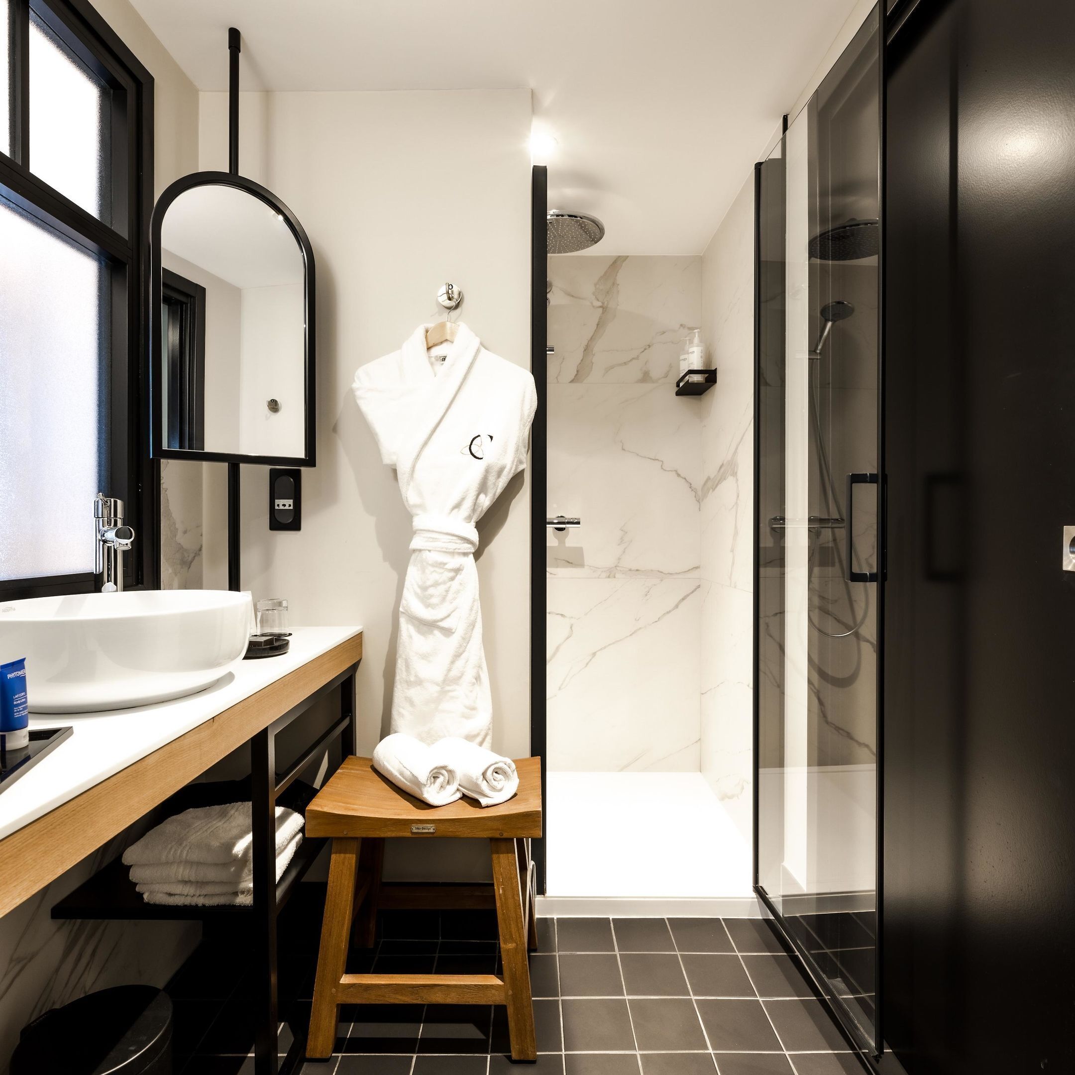 A modern bathroom with a white sink, a wooden stool with towels, a robe, and a shower area.