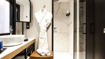 A modern bathroom with a white sink, a wooden stool with towels, a robe, and a shower area.