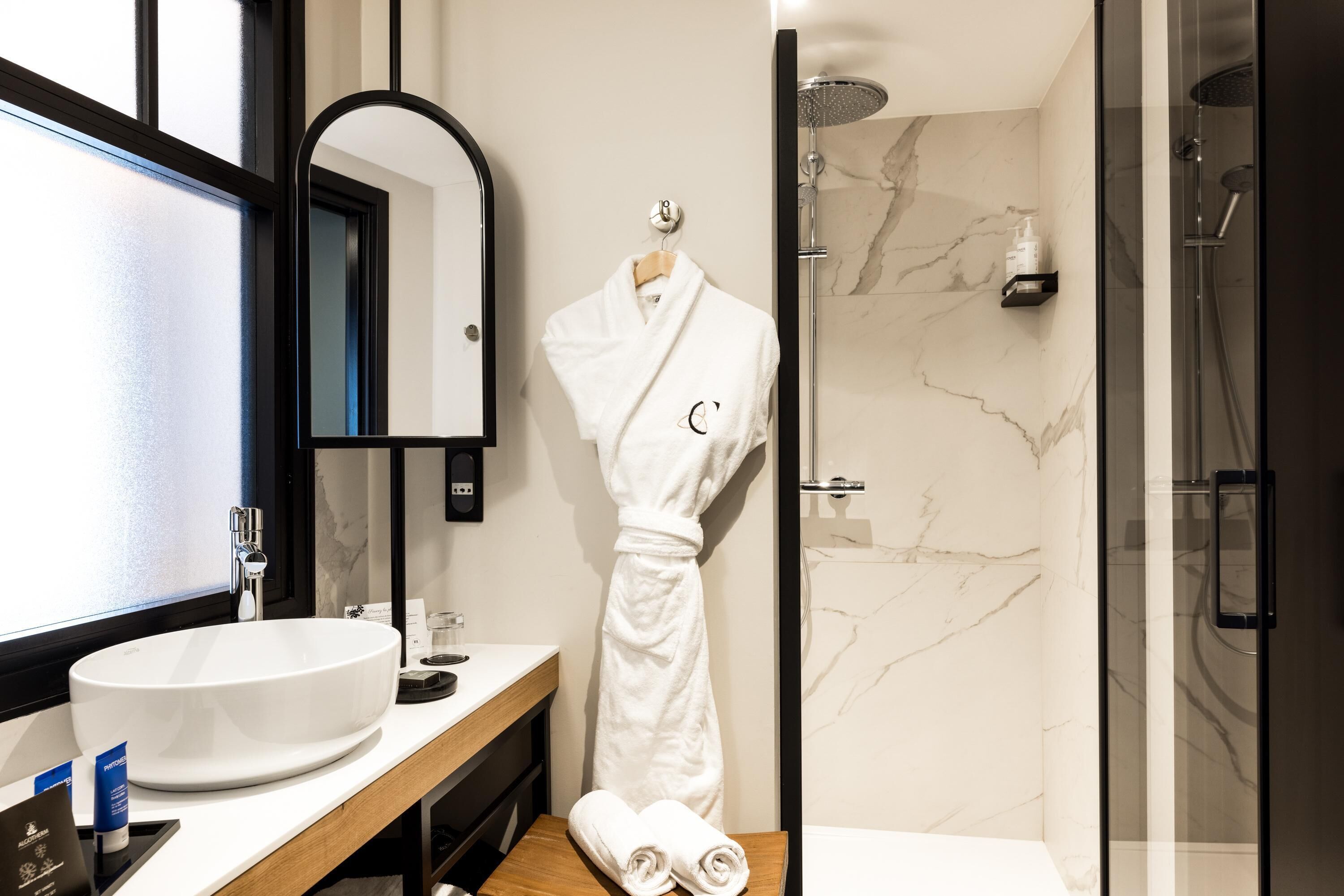 Un baño moderno con un lavamanos grande blanco, un espejo, una bata colgada en un gancho y una zona de ducha con mampara de vidrio.