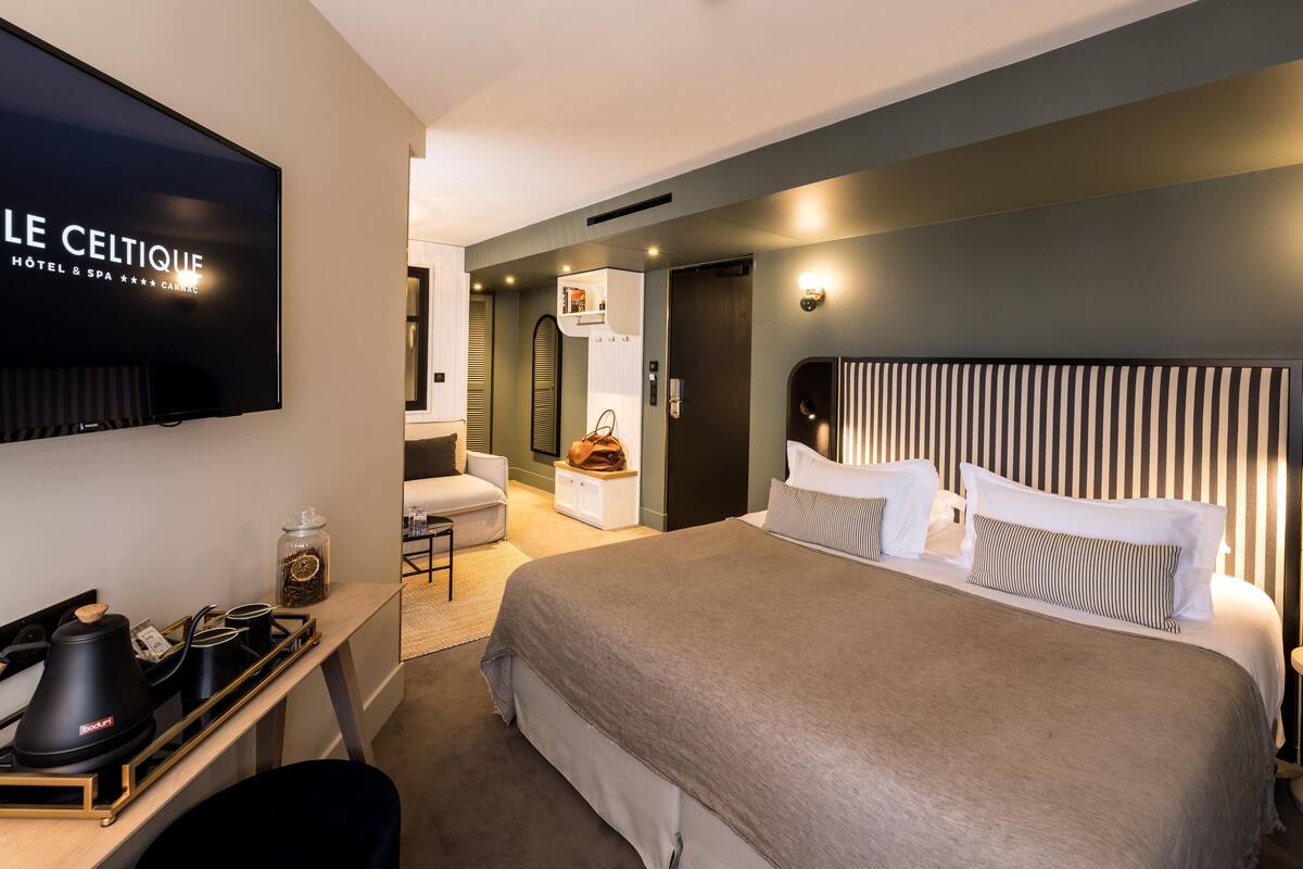 A modern hotel room with a large bed, a flat-screen TV, a desk with a coffee maker, and a small seating area.