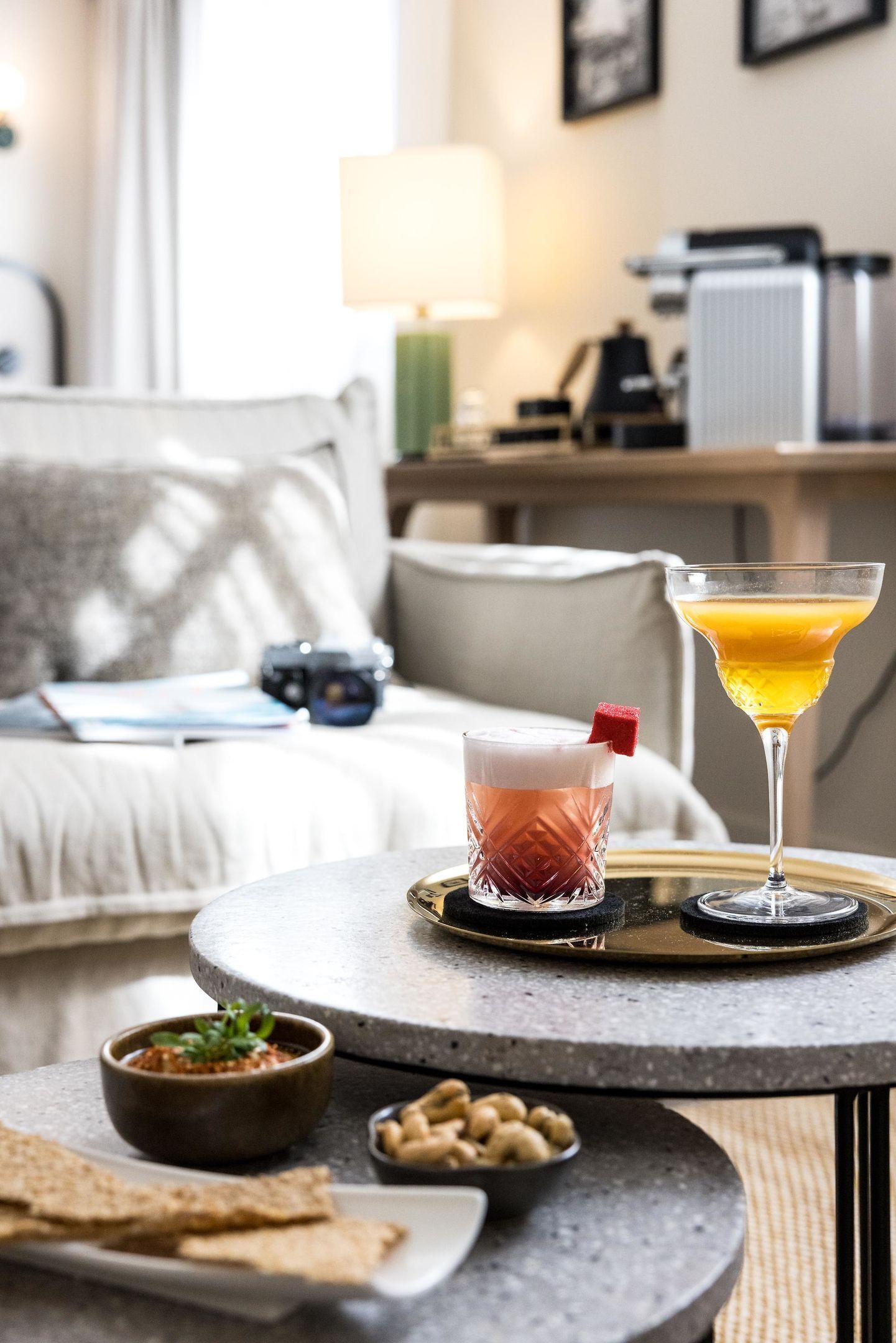 A round table with a martini glass and a glass of orange juice, a bowl of dip, and crackers on a tray.