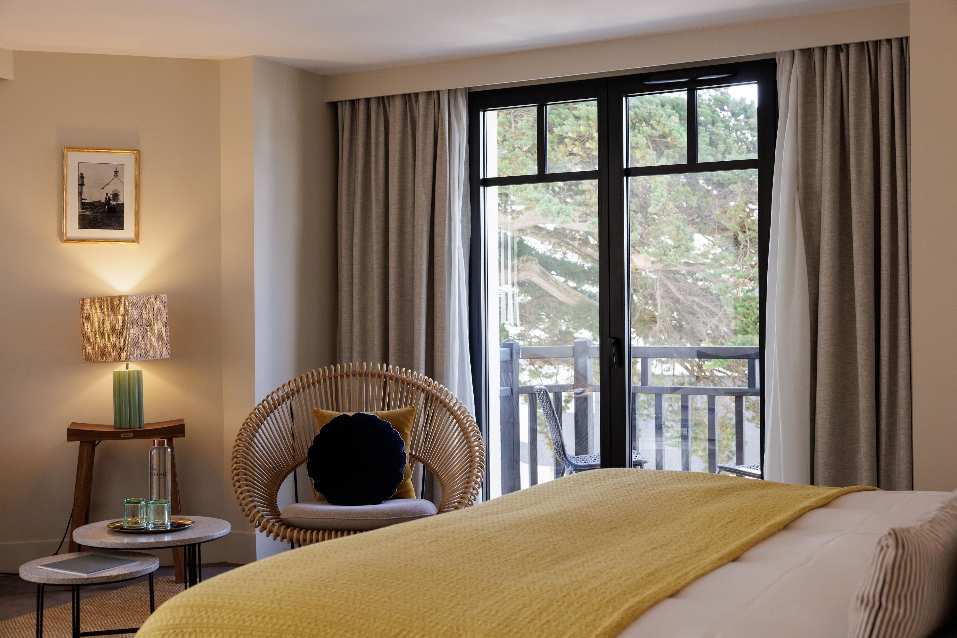A hotel room with a bed, a chair, a small table with a lamp, and a window with curtains.
