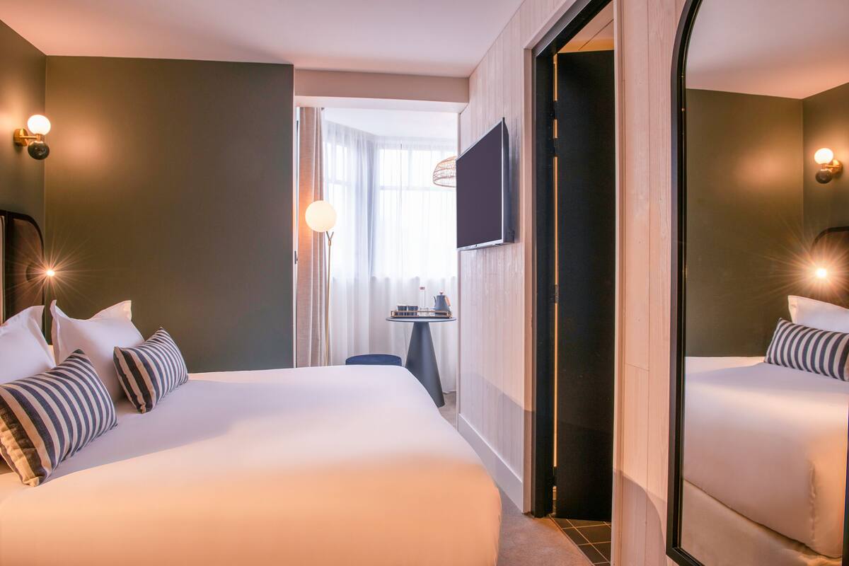 A hotel room with a large bed, a TV mounted on the wall, a small table with a lamp, and a mirror reflecting the bed.