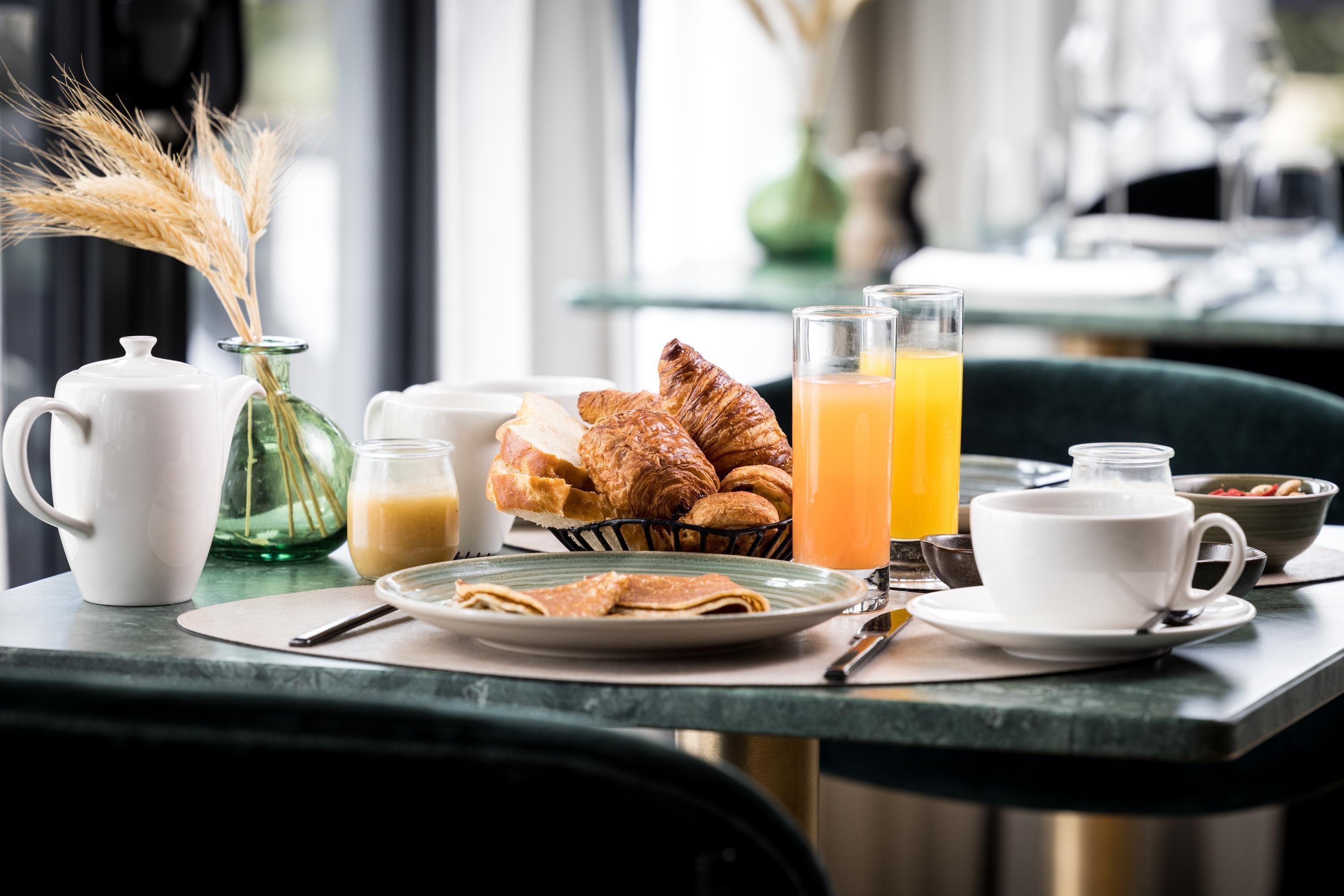 A breakfast table with croissants, a croissant basket, a glass of orange juice, a teapot, a cup, and a bowl of fruit.