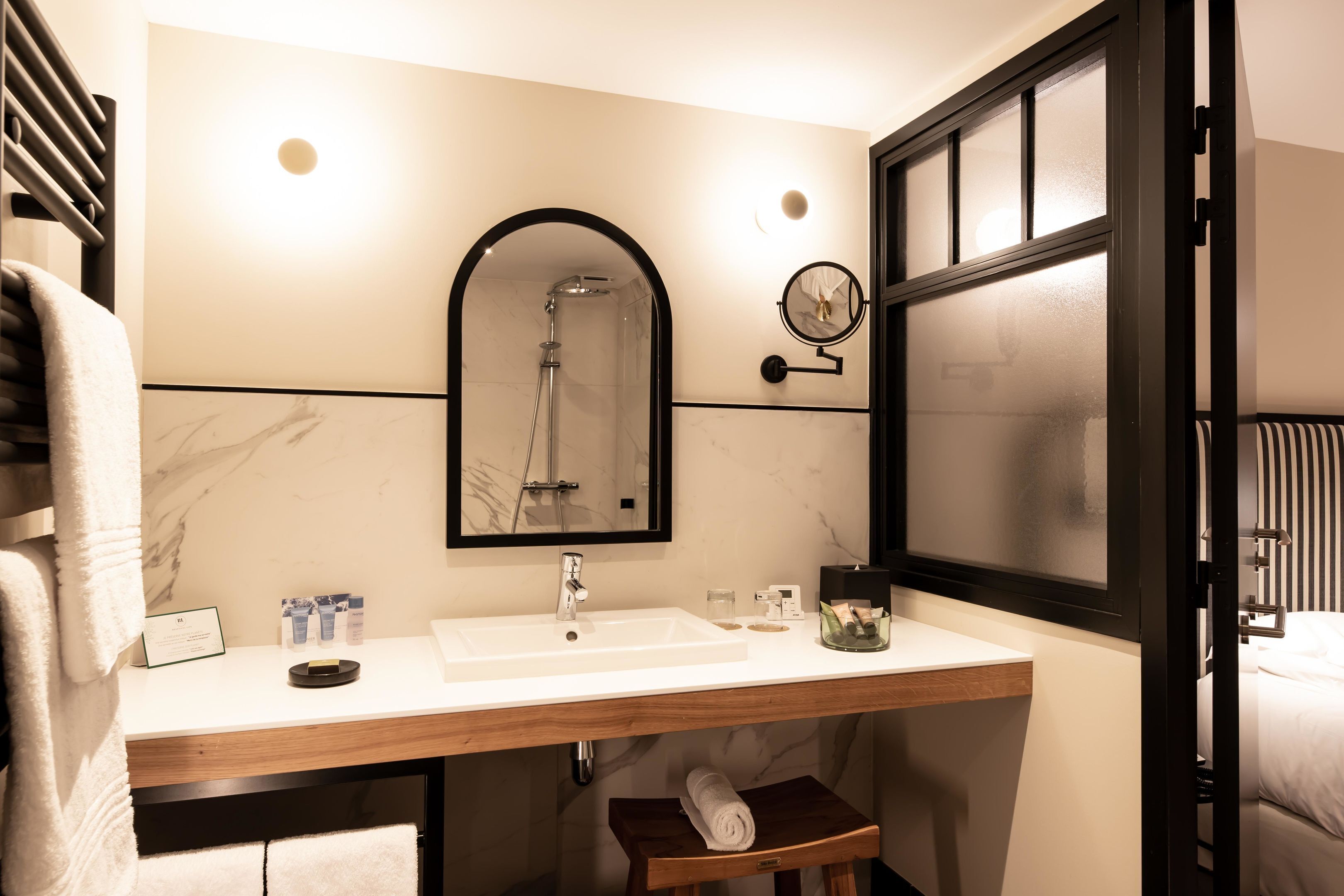 Un baño moderno con un lavabo blanco, un espejo y una ducha. Hay un perchero y un taburete de madera pequeño.