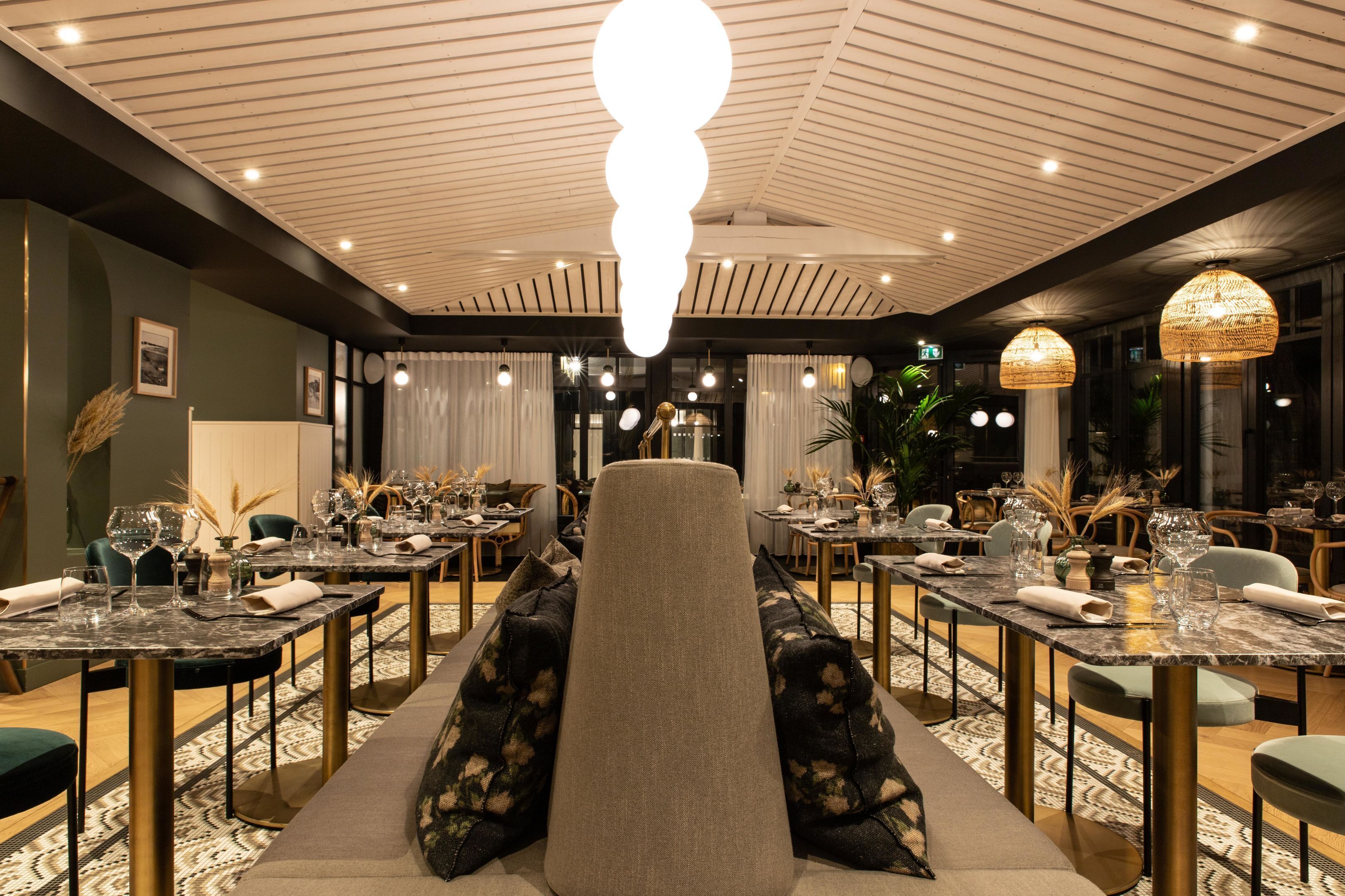 Restaurante com área de estar central, apresentando um amplo banco acolchoado. Mesas dispostas ao redor do banco, cada uma com copos e guardanapos. O teto possui um design único com nervuras e iluminação embutida.