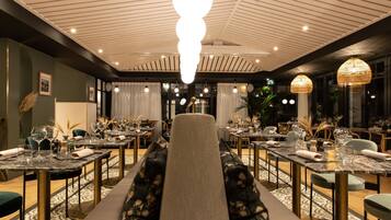 Restaurante com área de estar central, apresentando um amplo banco acolchoado. Mesas dispostas ao redor do banco, cada uma com copos e guardanapos. O teto possui um design único com nervuras e iluminação embutida.