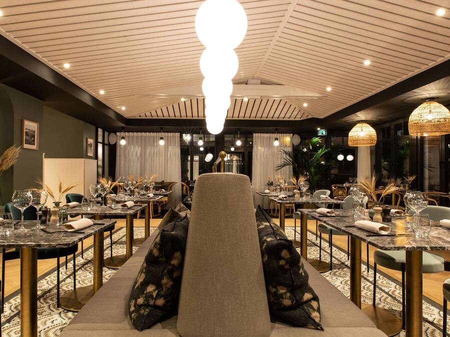 Un restaurante con una zona central de asientos que incluye un amplio banco acolchado. Mesas dispuestas alrededor del banco, cada una con copas y servilletas. El techo presenta un diseño único con nervaduras e iluminación empotrada.