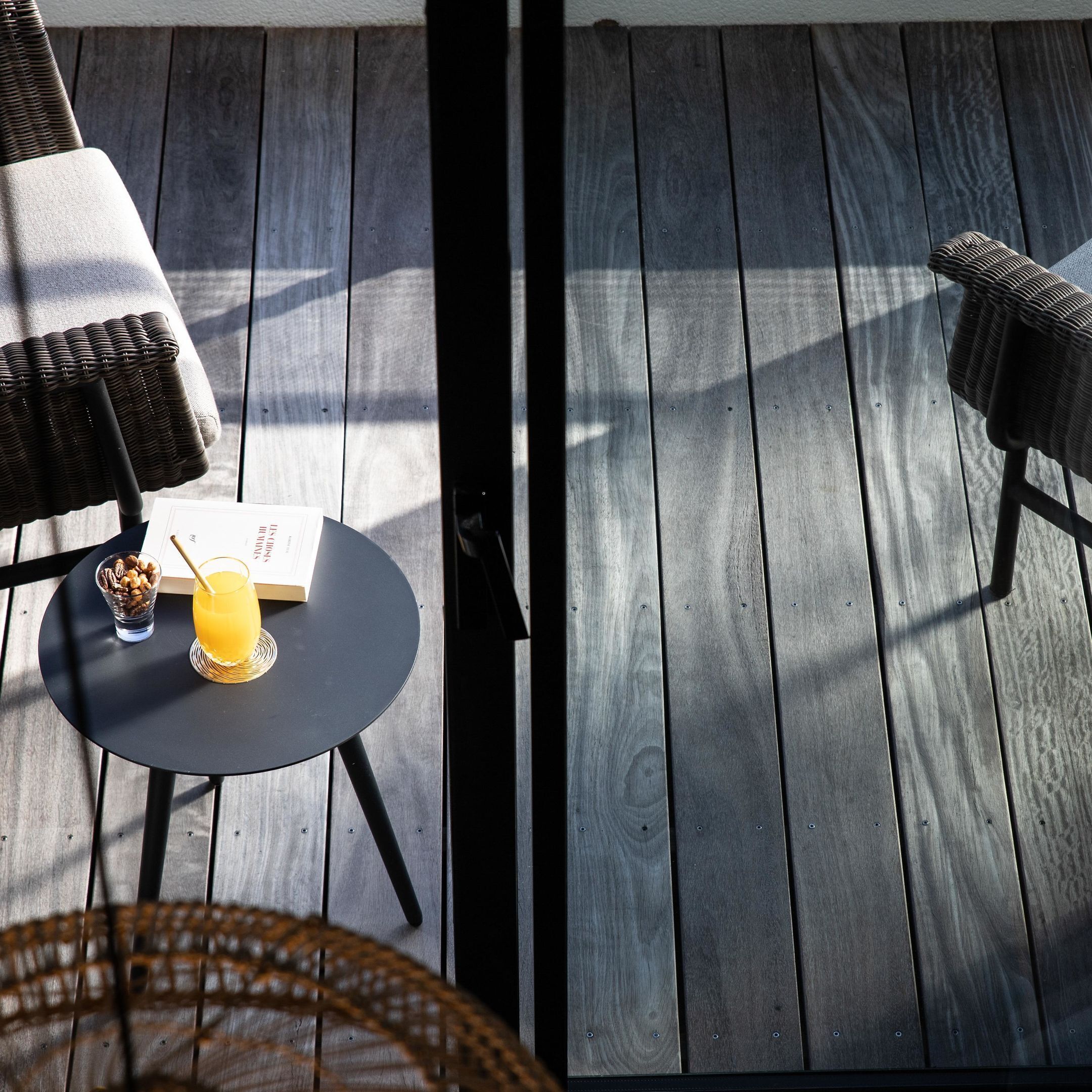 A wooden deck with two wicker chairs, a small round table with a drink and a book, and a wicker basket.
