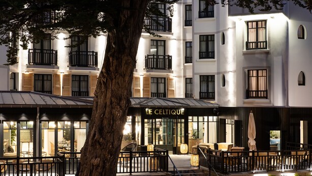 A modern hotel with a cafe area featuring outdoor seating and a large tree in the foreground. - Le Celtique & Spa (Carnac)