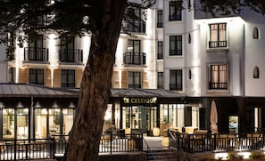 A modern hotel with a cafe area featuring outdoor seating and a large tree in the foreground.