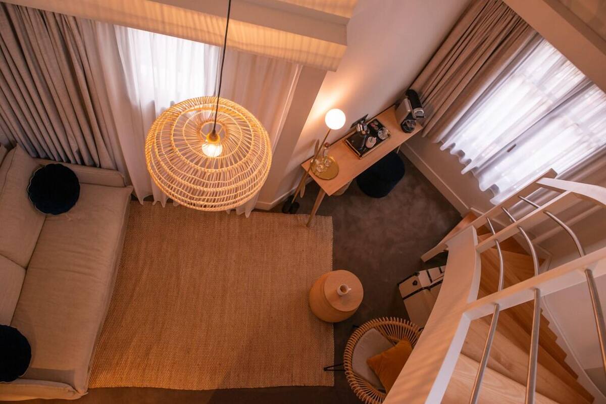 A modern hotel room with a large window, a woven light fixture, a sofa, a small table with a lamp, and a staircase.