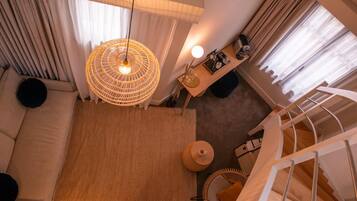 A modern hotel room with a large window, a woven light fixture, a sofa, a small table with a lamp, and a staircase.