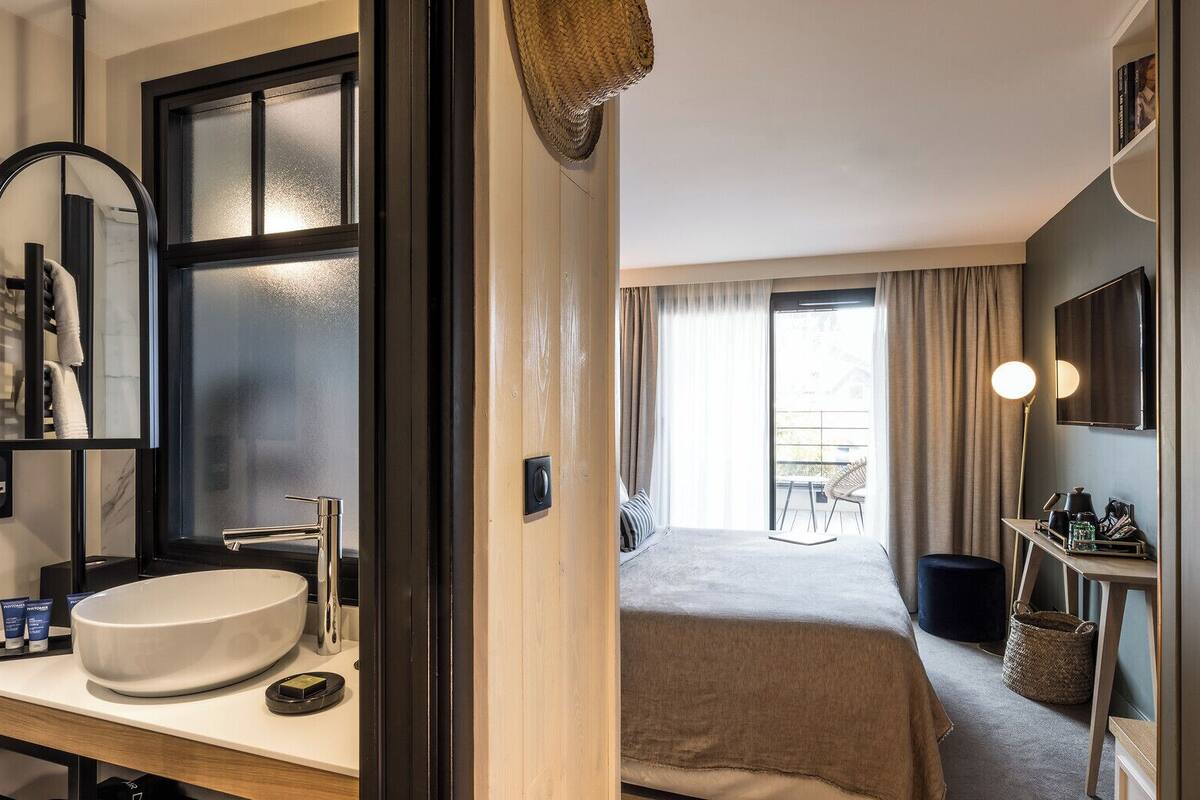 A modern hotel room with a large round sink, a bed with a beige blanket, a small table with a lamp, and a view of the outdoors through a large window.
