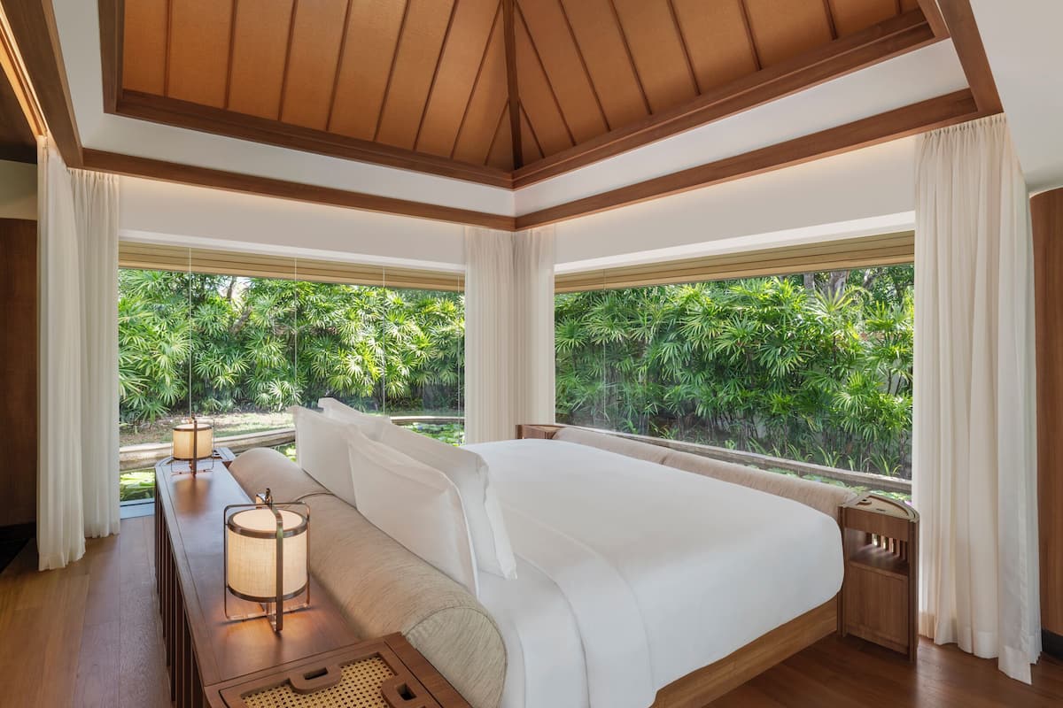 A spacious bedroom with a large bed, wooden headboard, and a view of lush greenery through large windows.
