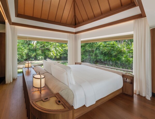 A spacious bedroom with a large bed, wooden headboard, and a view of lush greenery through large windows.