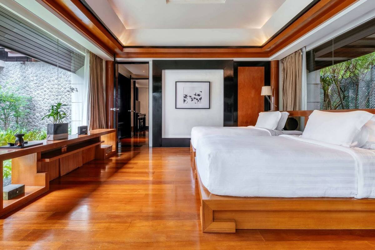 A modern hotel room with a wooden floor, a large bed, a desk, and a view of greenery outside.
