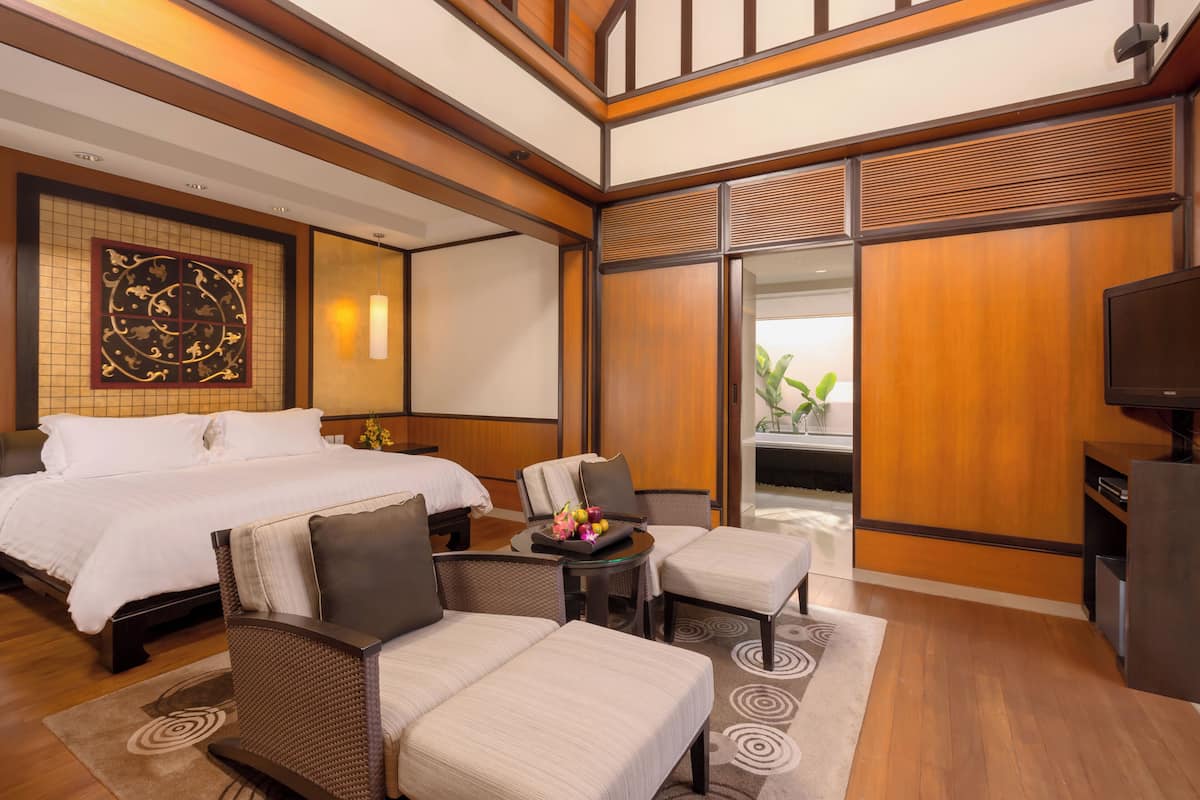 A hotel room with a large bed, two armchairs, a small table with fruit, a TV, and a view of a plant outside.