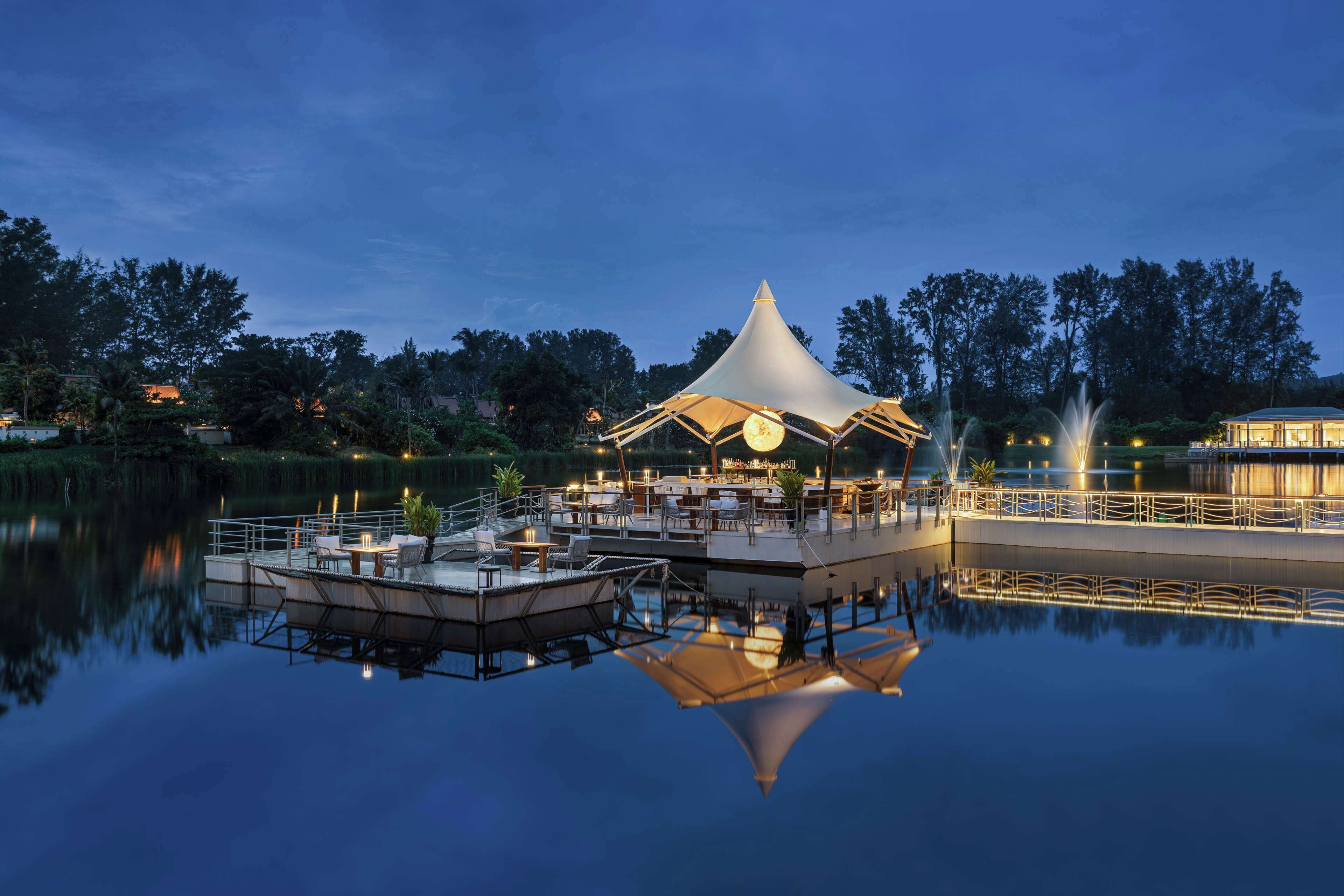 A tranquil lakeside setting with a dining area under a conical roof, surrounded by calm water and trees.