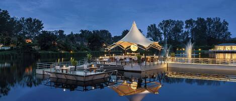 A tranquil lakeside setting with a dining area under a conical roof, surrounded by calm water and trees.