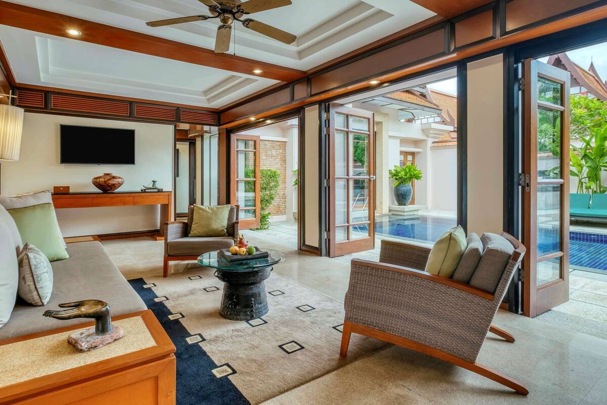 A spacious living area with a ceiling fan, a flat-screen TV, a sofa, a coffee table, and a view of an outdoor swimming pool.