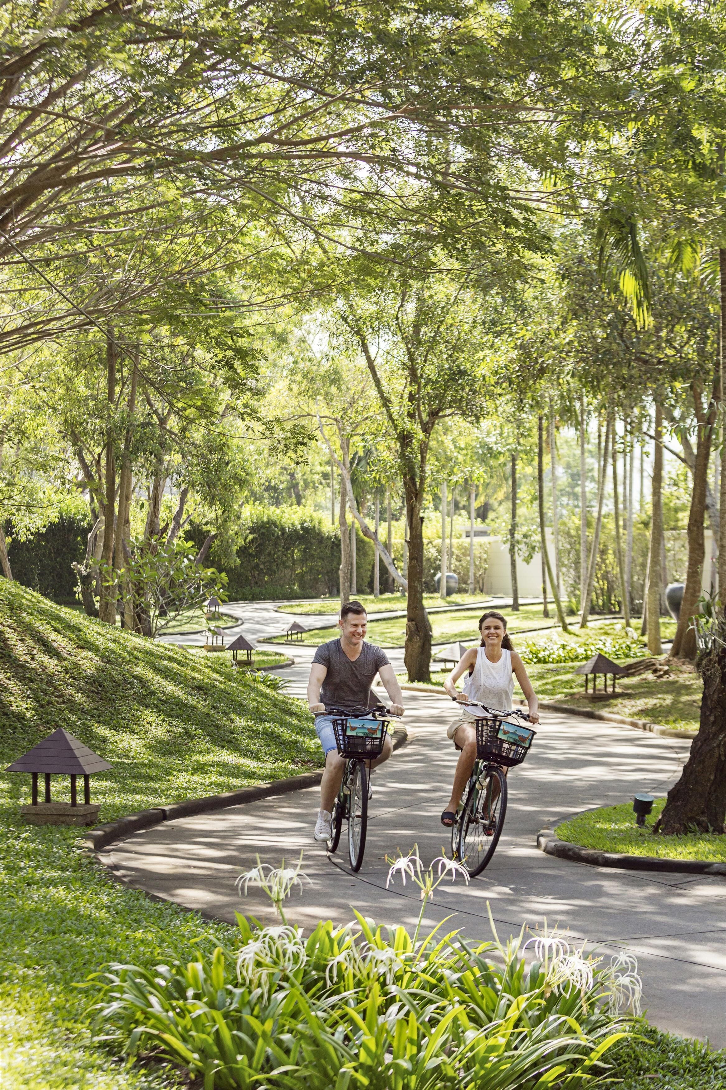 Dos personas montando en bicicleta por un camino pavimentado rodeado de exuberante vegetación y árboles.