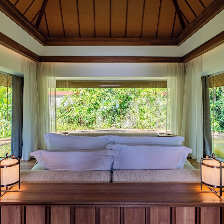 A bedroom with a large bed, two bedside lamps, and a view of a garden and pool.