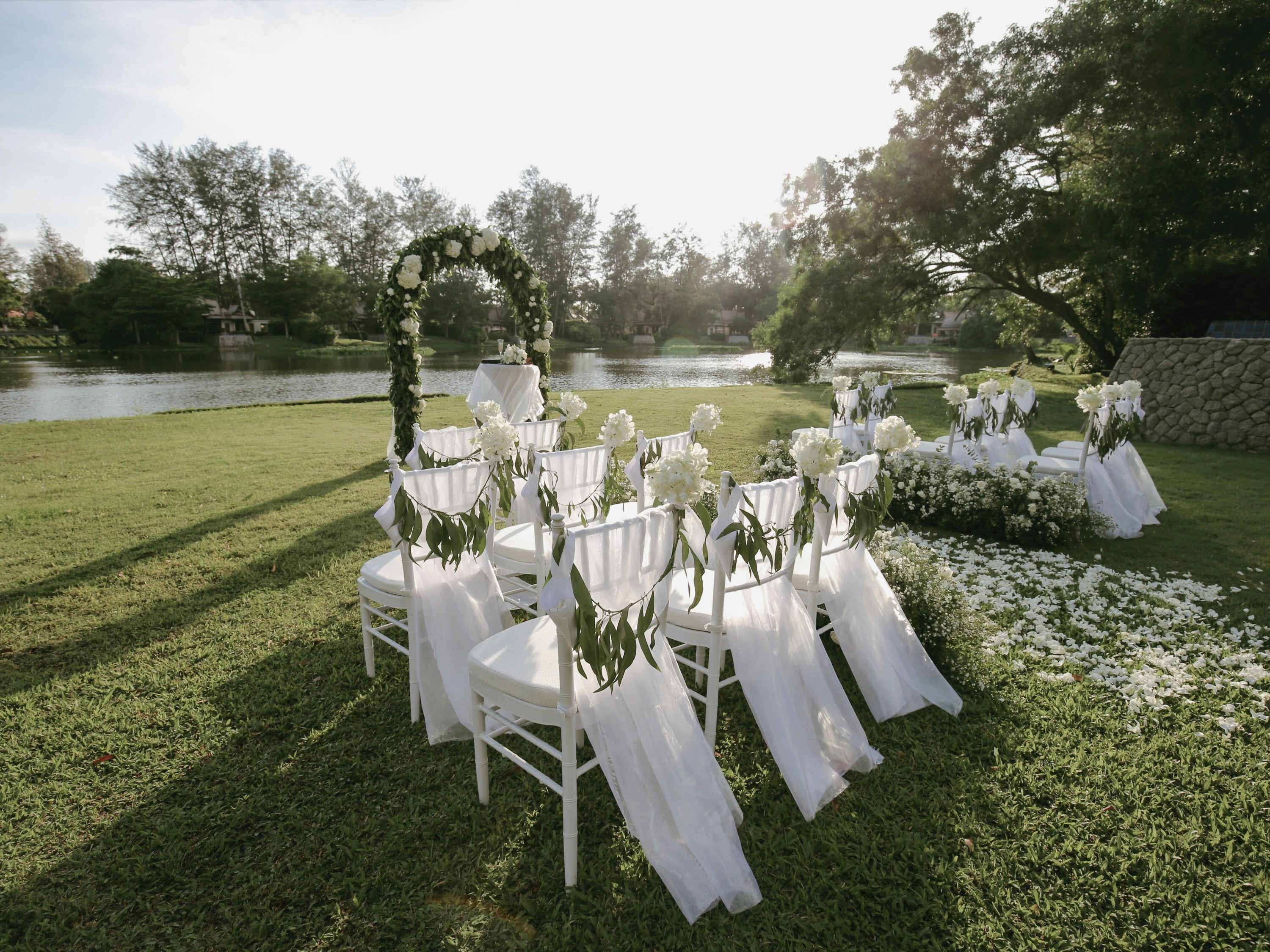 Decoración para una boda al aire libre con sillas y mesas blancas, un arco floral y un lago al fondo.