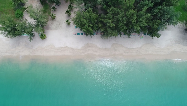 An aerial view of a beach with a row of beach huts along the shoreline, surrounded by dense greenery and palm trees. - Banyan Tree Phuket (Choeng Thale)
