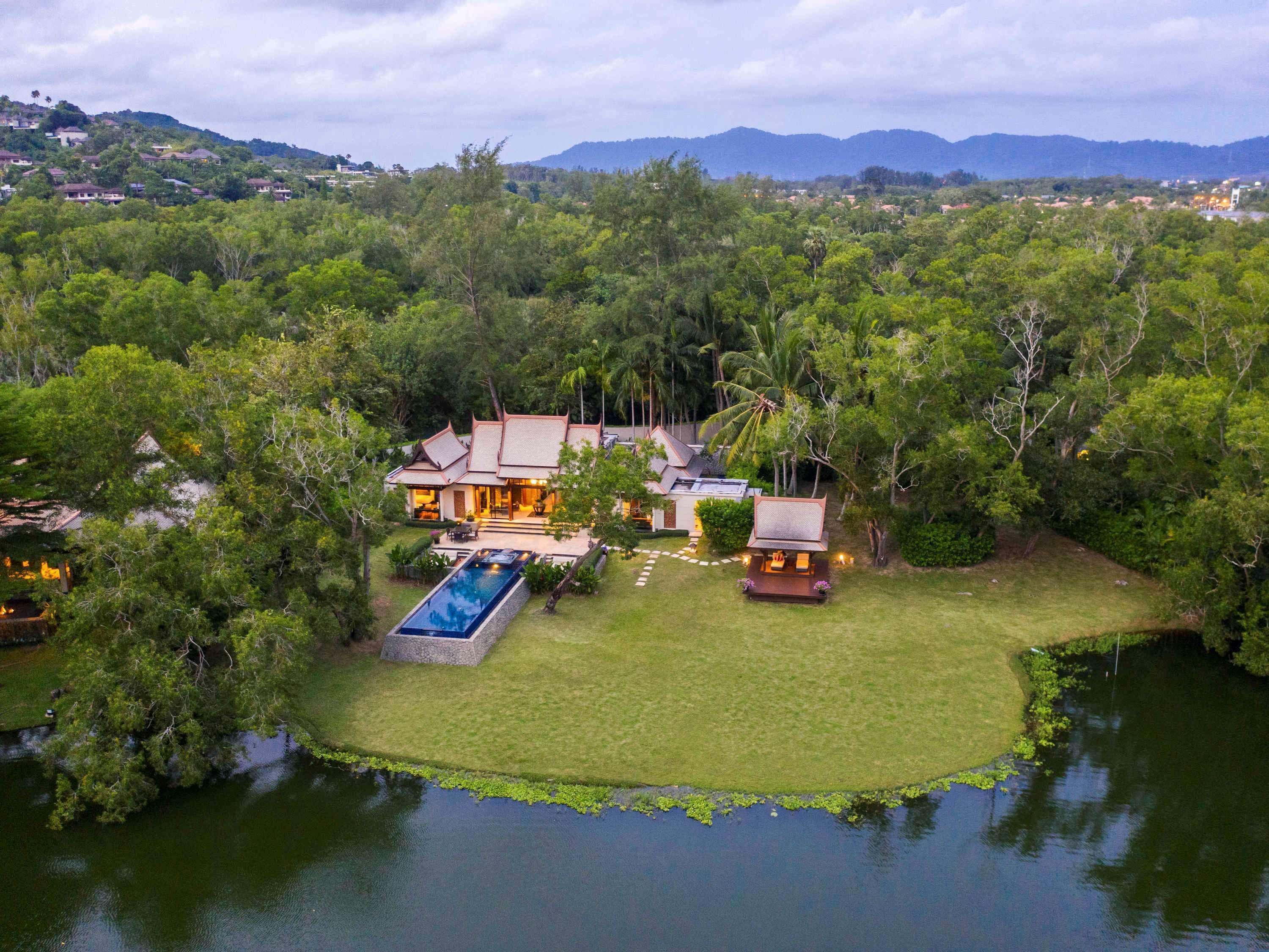 A large house with a swimming pool, surrounded by lush greenery and a lake.