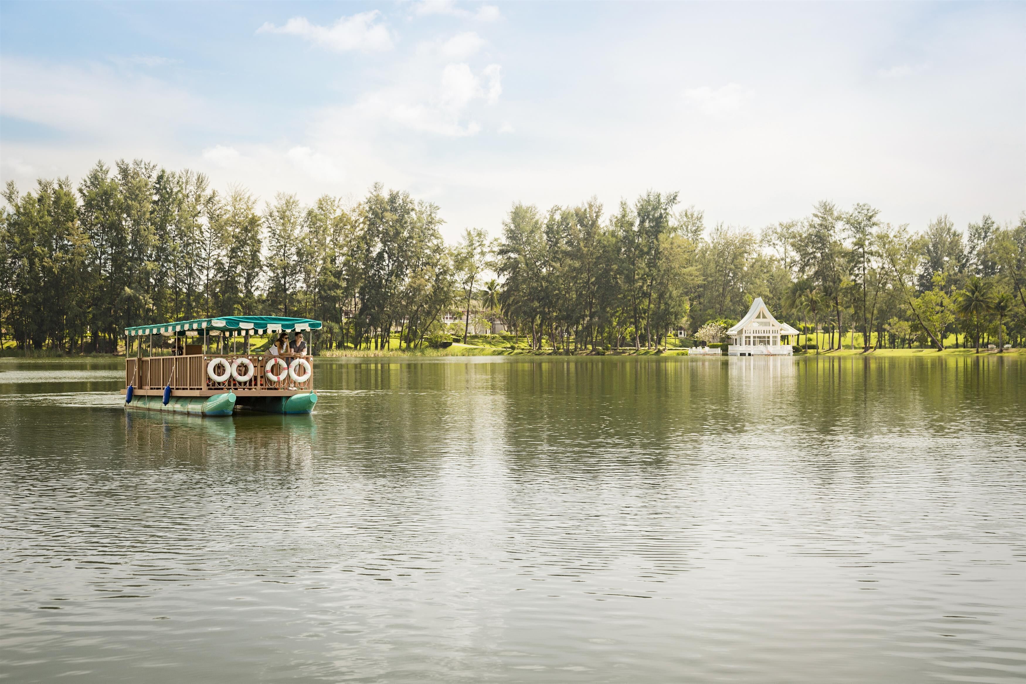 Un bote con toldo verde y boyas salvavidas en un lago tranquilo, rodeado de árboles y un pabellón blanco.