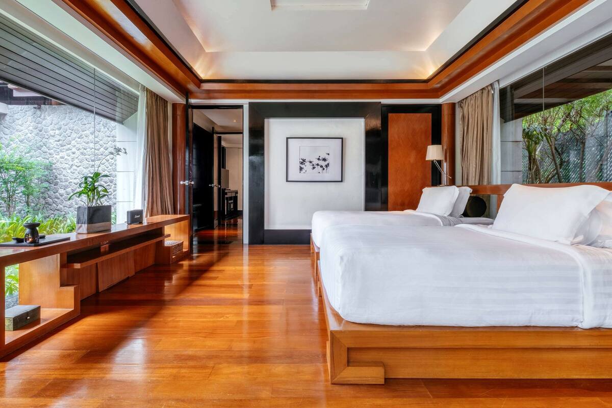 A modern hotel room with a wooden floor, a large bed, a desk, and a view of greenery outside.