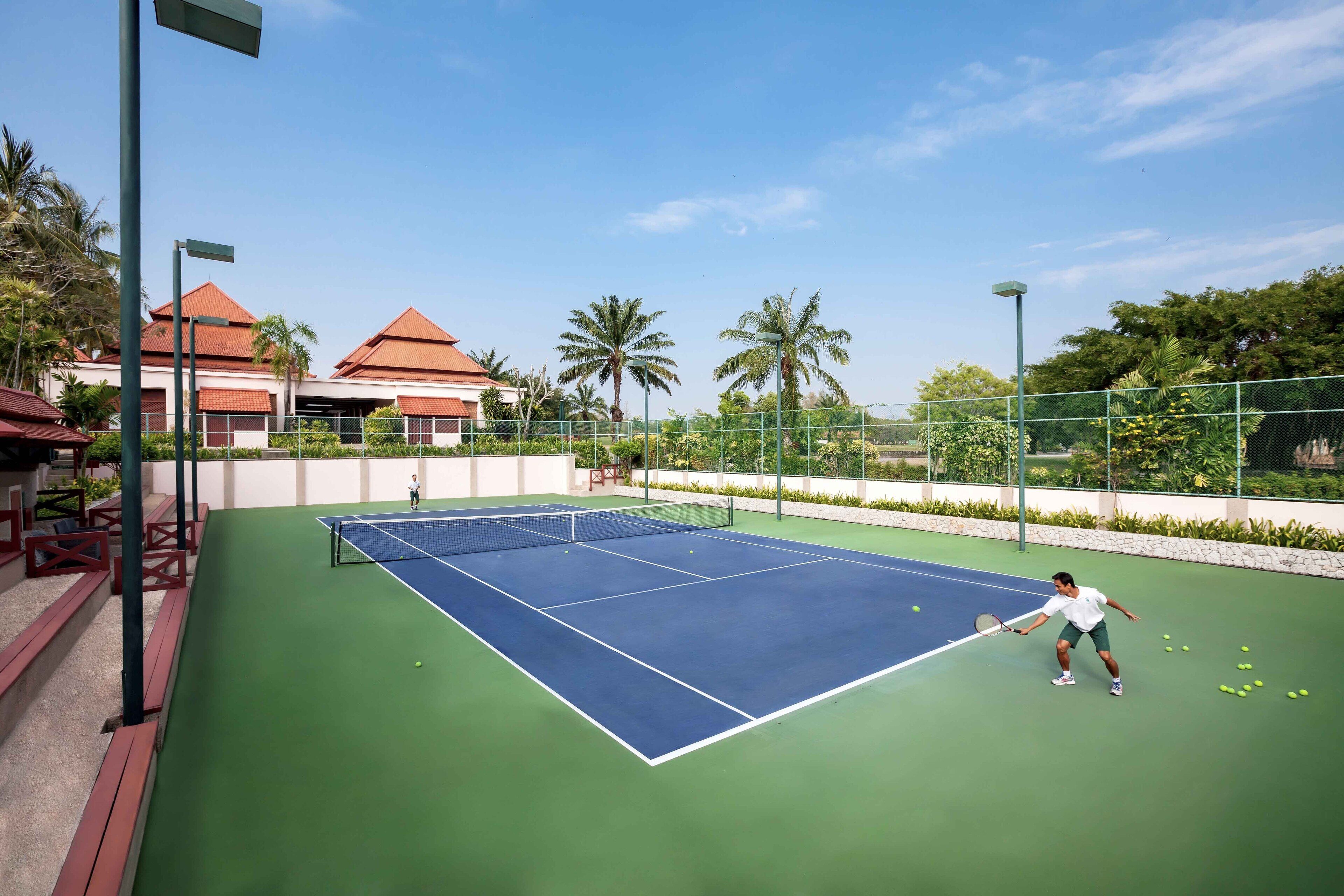 Ein Tennisplatz mit Netz, umgeben von Grünflächen und einem Gebäude mit rotem Dach.