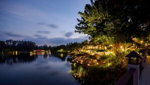 A lakeside hotel with a well-lit outdoor area, a bridge, and a reflection on the water.