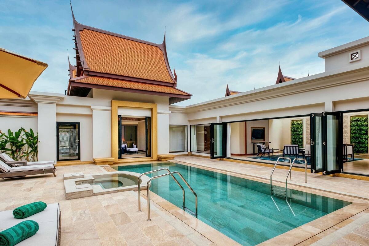 A modern hotel with a swimming pool, lounge chairs, and a traditional building with a tiled roof.