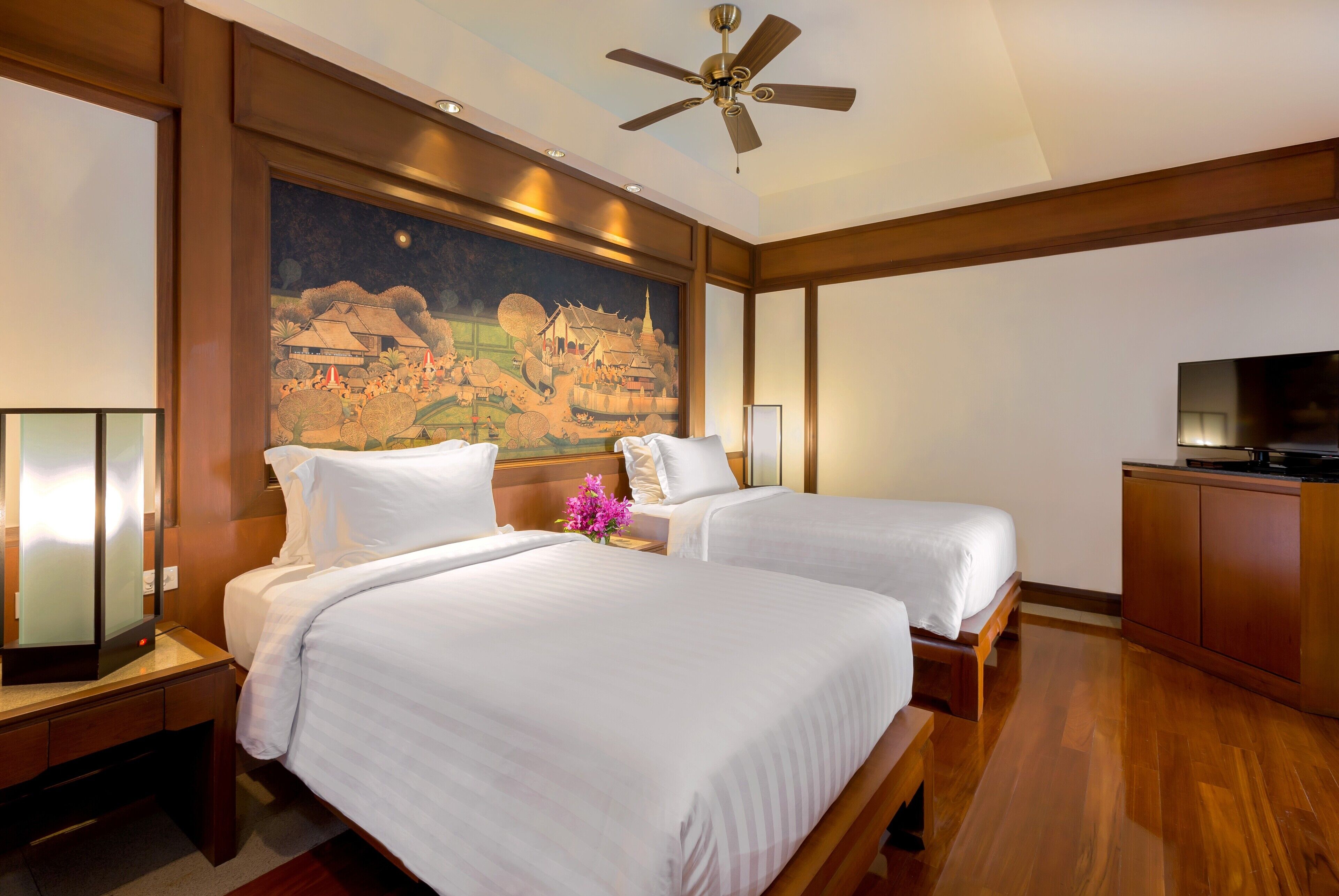 A hotel room with two beds, a ceiling fan, a painting on the wall, and a TV on a wooden cabinet.