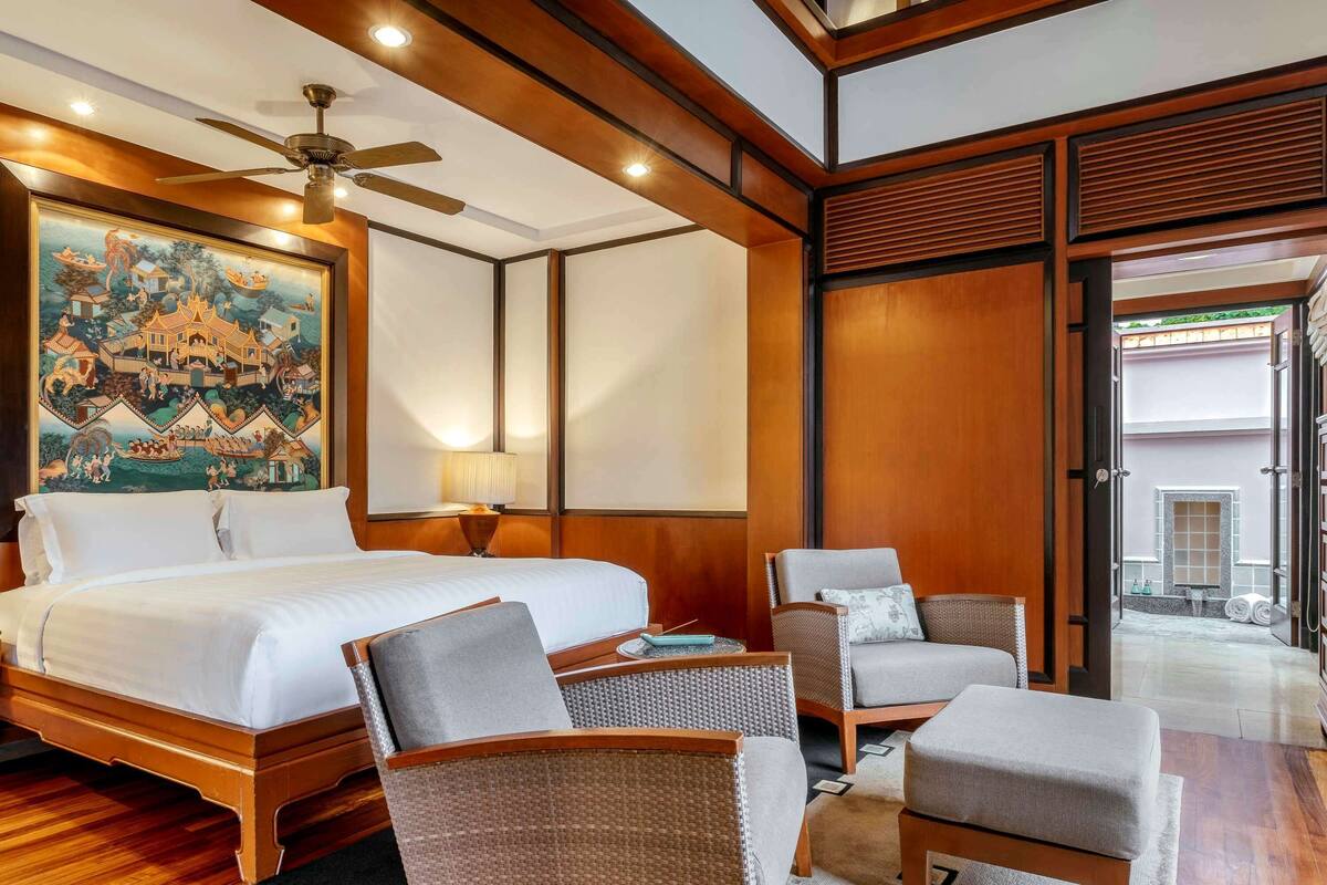 A hotel room with a large bed, a ceiling fan, a painting of a traditional scene, and a view of the outside through a glass door.