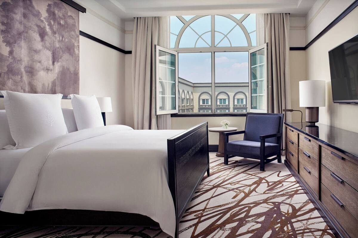 Habitación de hotel con una cama grande, una silla, un escritorio y un televisor. Se aprecia una ventana con vista a un edificio.