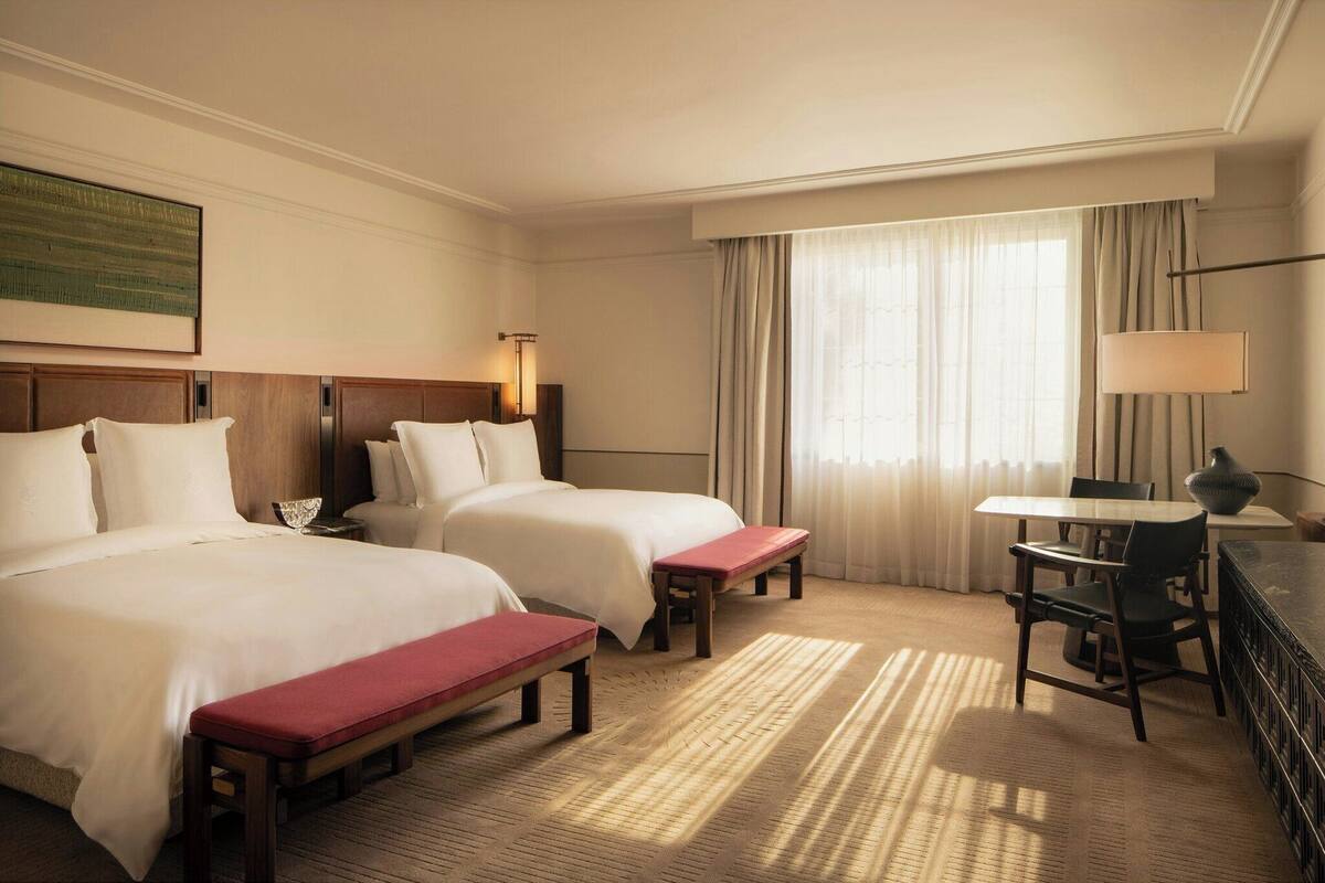 Habitación de hotel con dos camas, un escritorio, una silla, una lámpara y un ventanal grande con cortinas.